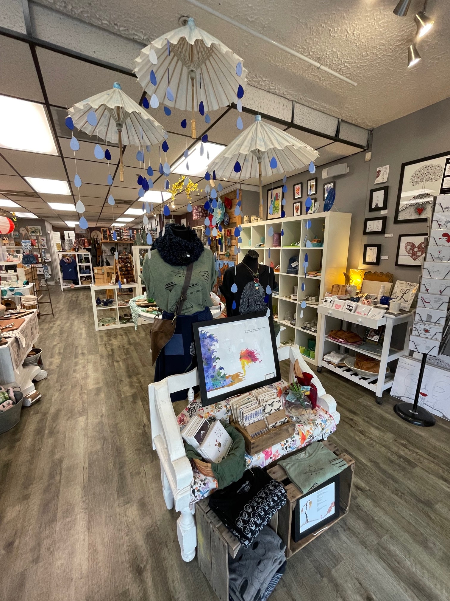 Image shows the interior of The Muse and a close up of a display featuring artwork, t-shirts and tea towels. Above the display are umbrellas and wood raindrops hanging from the ceiling. 